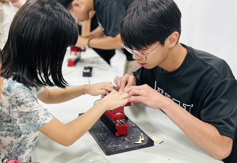 Skilled instructor Guan-Wei Liu (right) guides children in Nantou in creating laser-engraved wooden music boxes.